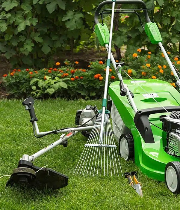 A green lawn mower alongside gardening tools rests on a lush green lawn, indicating yard work preparation.
